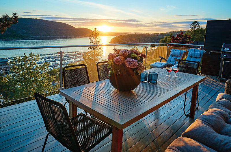 Terrasse mit Holz-Gartentisch und Stühlen, 2 Weingläser, Ausblick auf einen See mit Sonnenuntergang